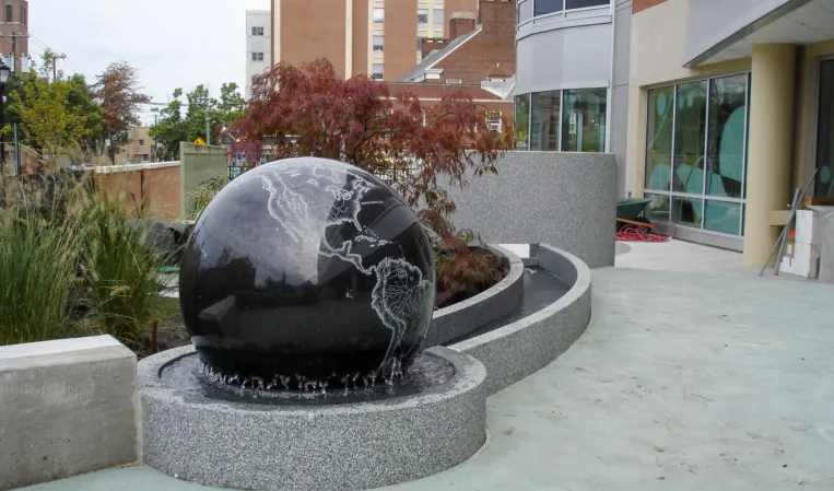 Polished black granite sphere fountain with a curved water channel in a landscaped urban setting