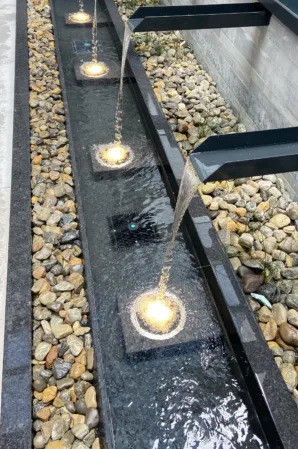 Illuminated water jets flowing into a narrow granite basin lined with pebbles