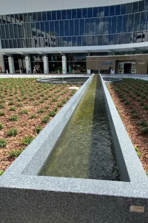 Long, narrow granite water channel bordered by landscaped planting beds outside a modern glass building