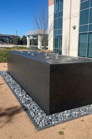 Black granite fountain with bubbling water jets in a landscaped outdoor area beside a modern building