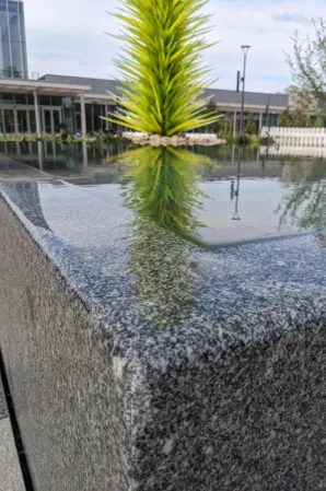 Close-up of the polished granite edge of a reflecting pool with a green glass sculpture in the background