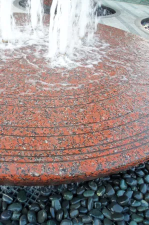 Close-up of water jets splashing onto a textured red granite fountain surface surrounded by black pebbles