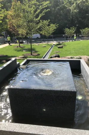 Square granite fountain with bubbling water and a narrow channel extending through a landscaped park