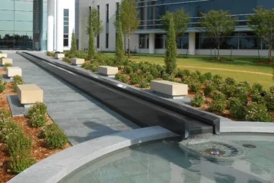Long, narrow granite water channel flowing into a circular fountain basin in a landscaped courtyard