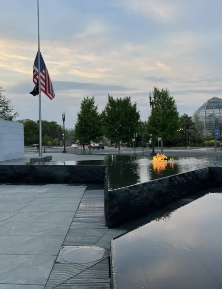 Granite reflecting pool with an illuminated sculpture and American flag at half-mast in a landscaped memorial plaza
