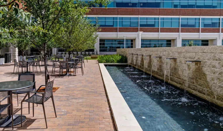 Stone fountain wall with water spouts beside an outdoor seating area and trees