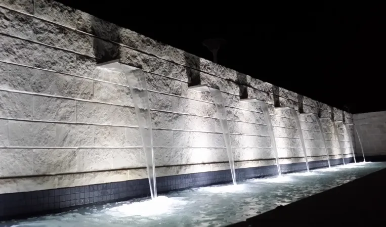 Illuminated stone wall with evenly spaced water spouts cascading into a long basin