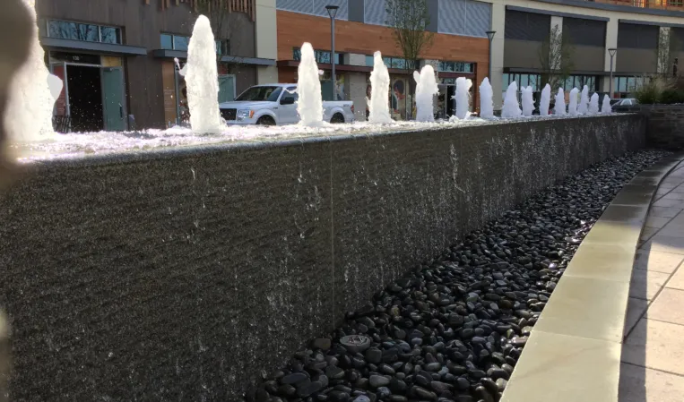 Row of water jets along a raised stone fountain wall with black pebbles below