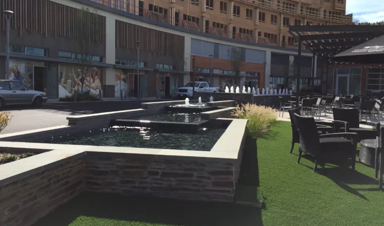 Raised stone fountain with water jets beside an outdoor dining area in an urban plaza