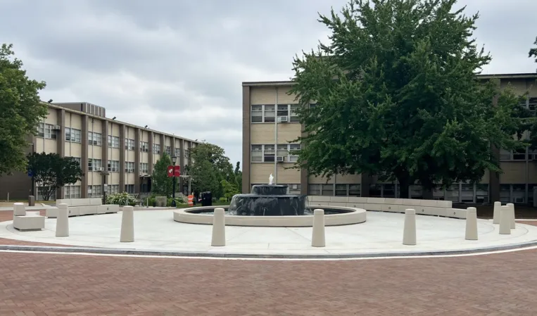 Circular stone fountain in a paved plaza surrounded by buildings and trees