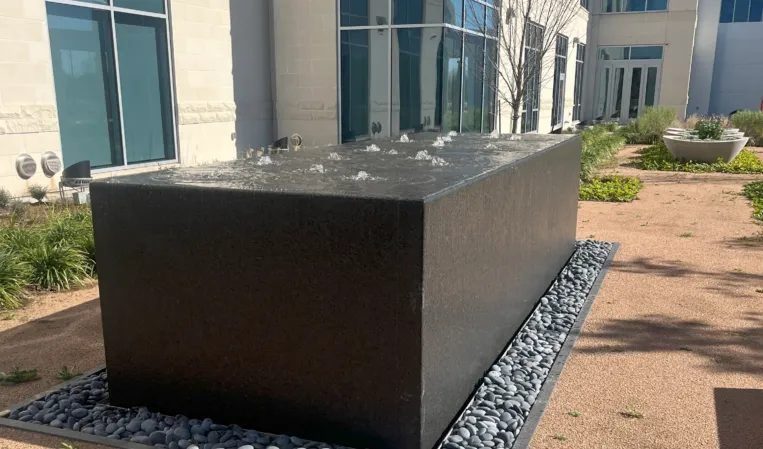 Black granite fountain with bubbling water jets in a landscaped outdoor area beside a modern building