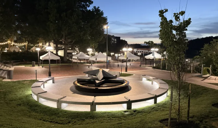 Illuminated circular granite memorial fountain with a central stone sculpture in a landscaped public plaza at dusk