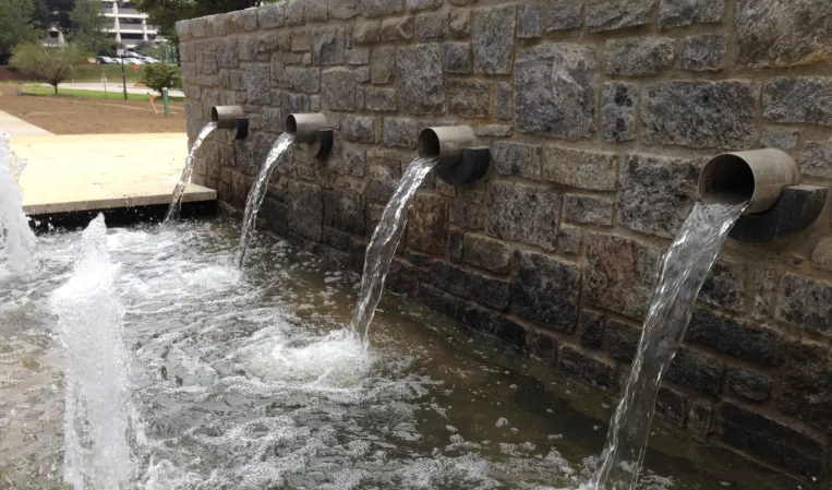 Close-up of four metal water spouts pouring into a fountain basin from a natural stone wall