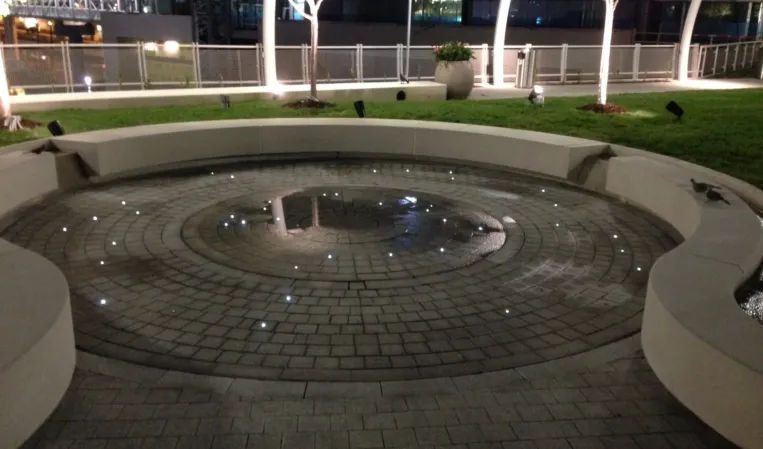 Circular fountain with embedded lights and concentric paving patterns in an illuminated landscaped plaza at night