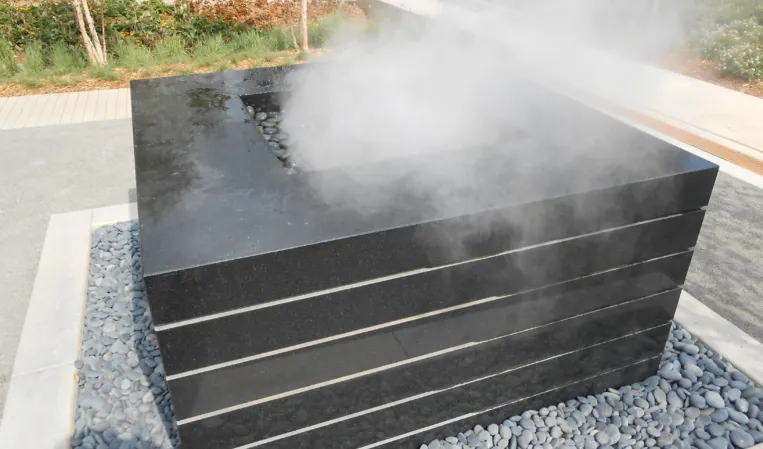 Rectangular black granite fountain with horizontal grooves and rising water mist, surrounded by grey pebbles