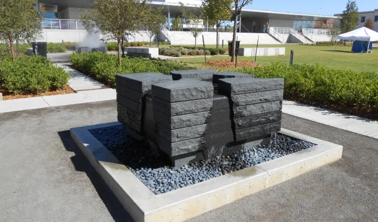 Sculptural fountain made of stacked granite blocks with water flowing into a square pebble-filled basin