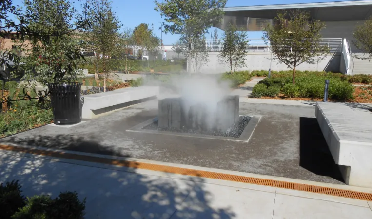 Granite fountain with rising water mist in a landscaped courtyard surrounded by trees and modern architecture