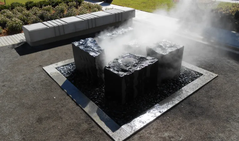 Three solid granite blocks surrounded by rising water mist in a square fountain basin in a public space