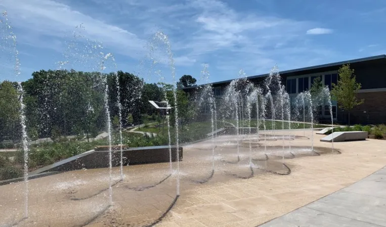 Interactive fountain with arched water jets in a paved plaza surrounded by greenery and modern buildings
