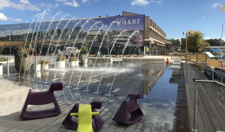 Interactive fountain with arched water jets over a paved plaza near a modern waterfront building