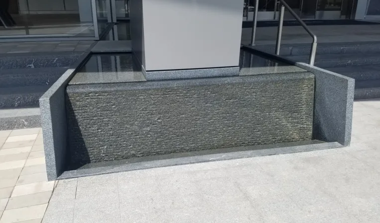 Textured granite water wall with a shallow reflecting pool beside a modern building entrance