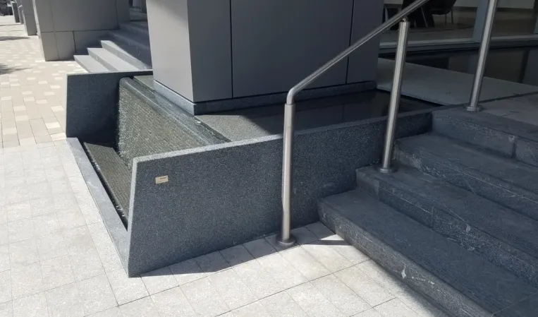 Textured granite water wall with a shallow reflecting pool beside a modern building entrance