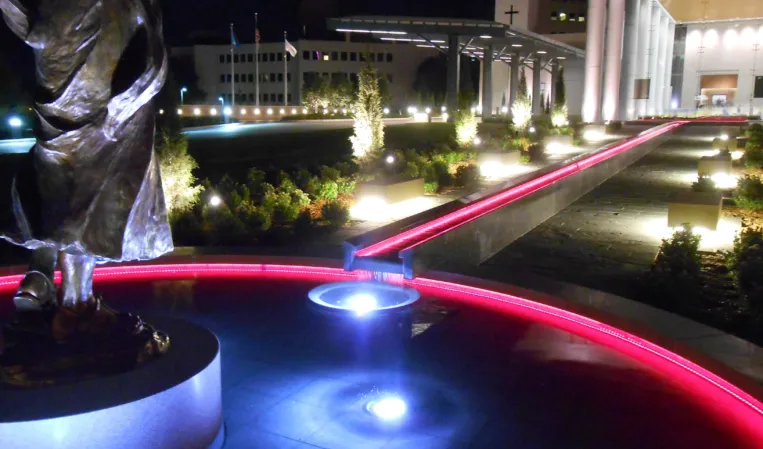 Illuminated fountain with a bronze sculpture, colorful lighting and a long water channel in a landscaped plaza at night