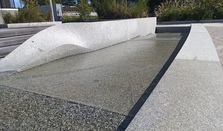 Close-up of a granite water feature with a sloped overflow edge and shallow basin beside a landscaped garden