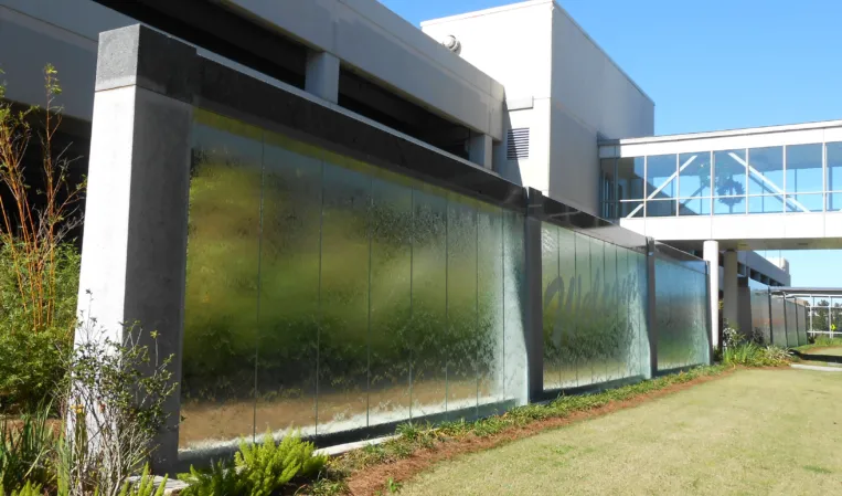 Glass water wall with cascading water beside a landscaped lawn and modern buildings