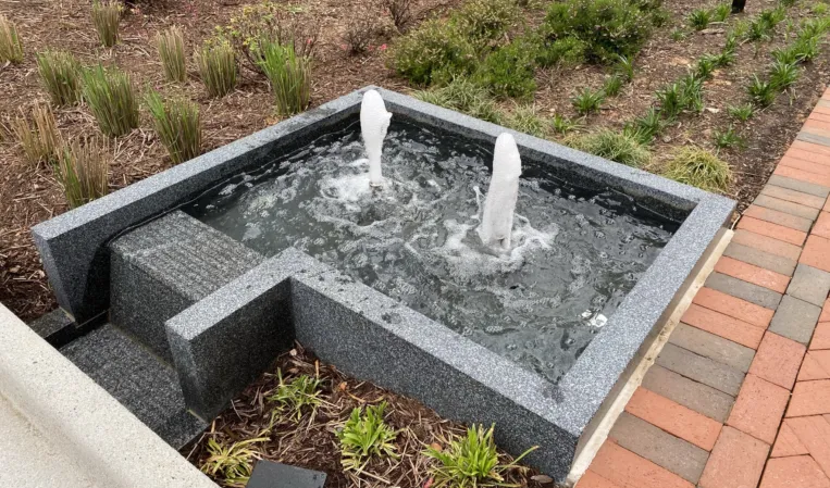 Square granite fountain with two bubbling water jets and a stepped water channel in a landscaped courtyard