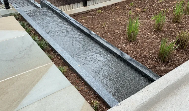 Narrow granite water channel with gently flowing water beside a landscaped planting bed