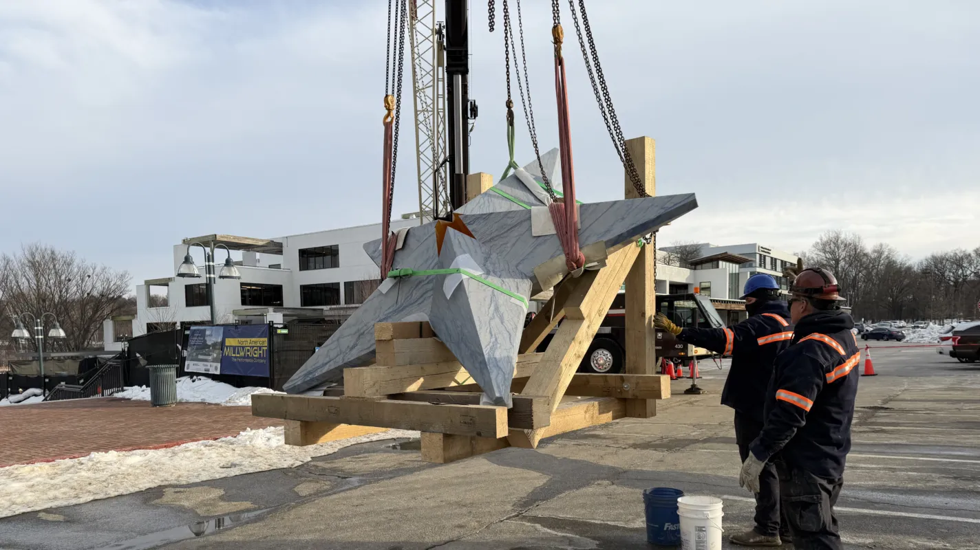 Workers guiding an angular stone sculpture into position with a crane at a construction site.