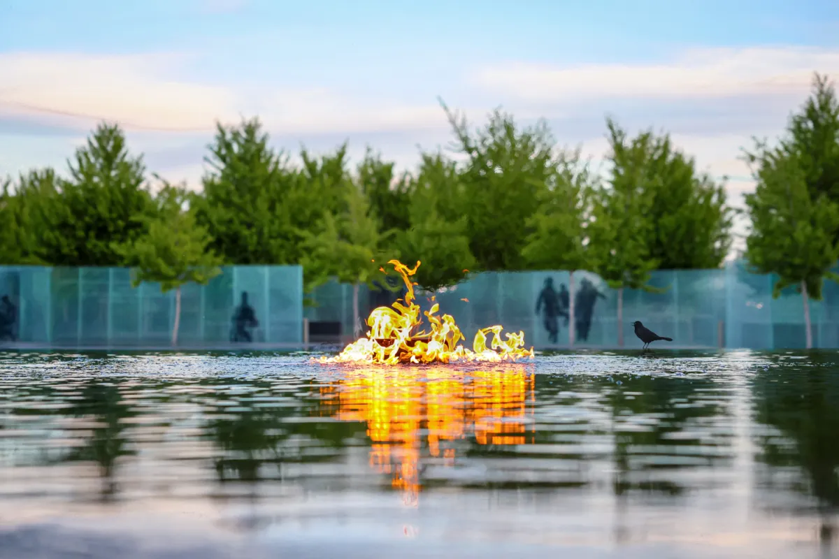Flames rising from a shallow reflecting pool, their orange glow mirrored in the water, with trees, translucent glass panels and a black bird in the background.