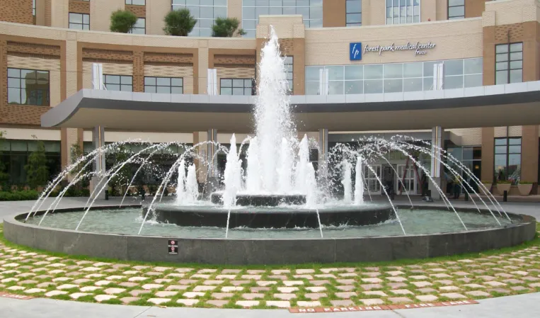 Circular fountain with a tall central water jet and arched jets outside a modern building