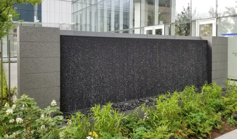 Large black granite water wall with cascading water, surrounded by greenery in front of a modern glass building