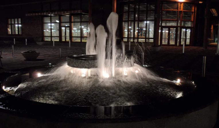 Circular granite fountain with multiple water jets cascading into a lower basin in an urban plaza at night